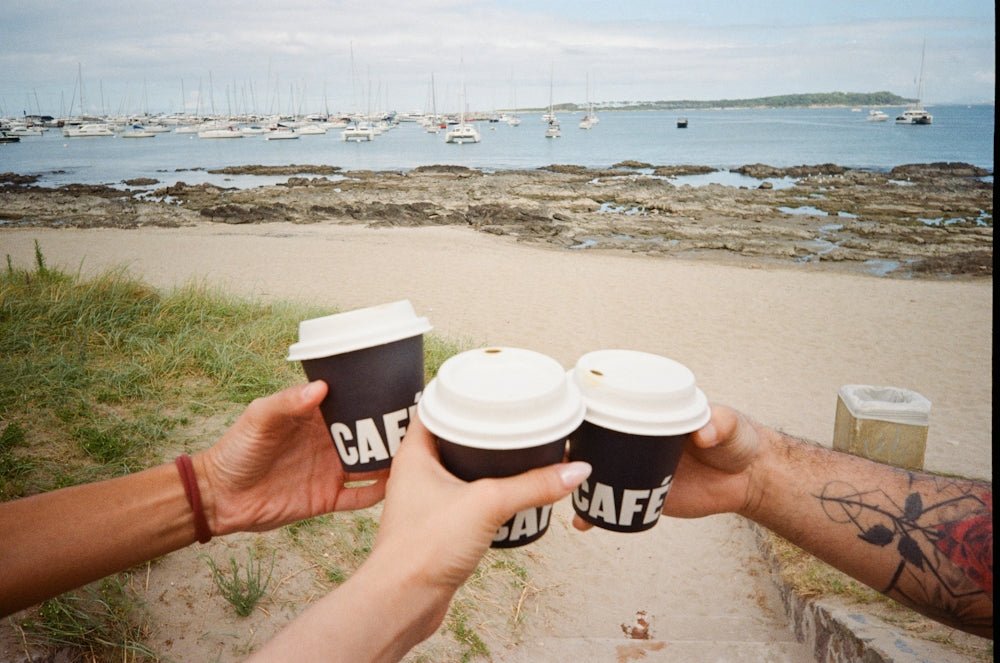 Three hands toast coffee cups on a beach with boats.