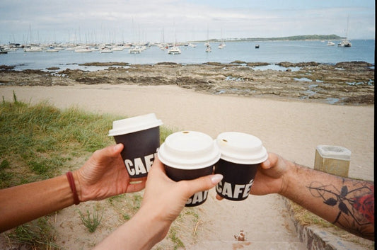 Three hands toast coffee cups on a beach with boats.